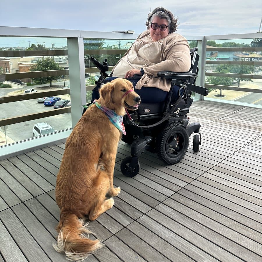 Kristen out on a rooftop deck in her power chair with Millie, her golden retriever, sitting calmly beside the wheel